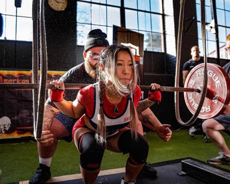 A lifter hits depth on a squat attempt with the spotter tracking the bar behind her.